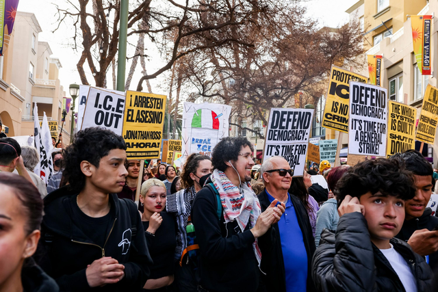 A diverse crowd marches through a city street holding signs demanding ICE removal, defending democracy, and calling for justice and a general strike.